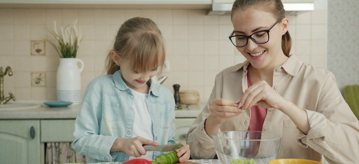 Helpful daughter is making vegetable salad chopping cucumber while young loving mother is touching her hair expressing love at home in kitchen.