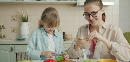 Helpful daughter is making vegetable salad chopping cucumber while young loving mother is touching her hair expressing love at home in kitchen.