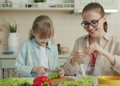 Helpful daughter is making vegetable salad chopping cucumber while young loving mother is touching her hair expressing love at home in kitchen.